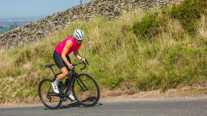 A female cyclist climbs out of the saddle in a pink top and white helmet with greenary in the background